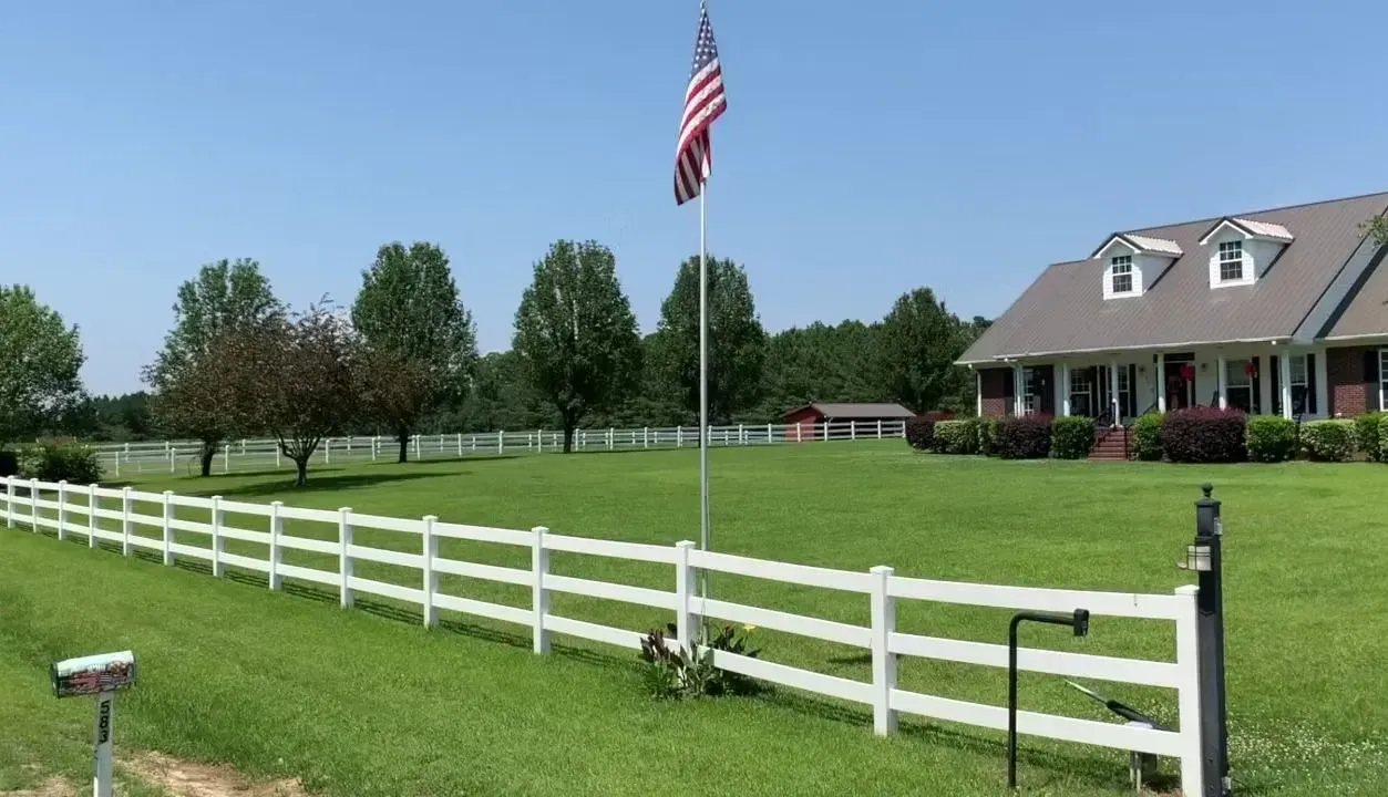 American flag on a tall pole in front of a white fence and a large house with a green lawn.