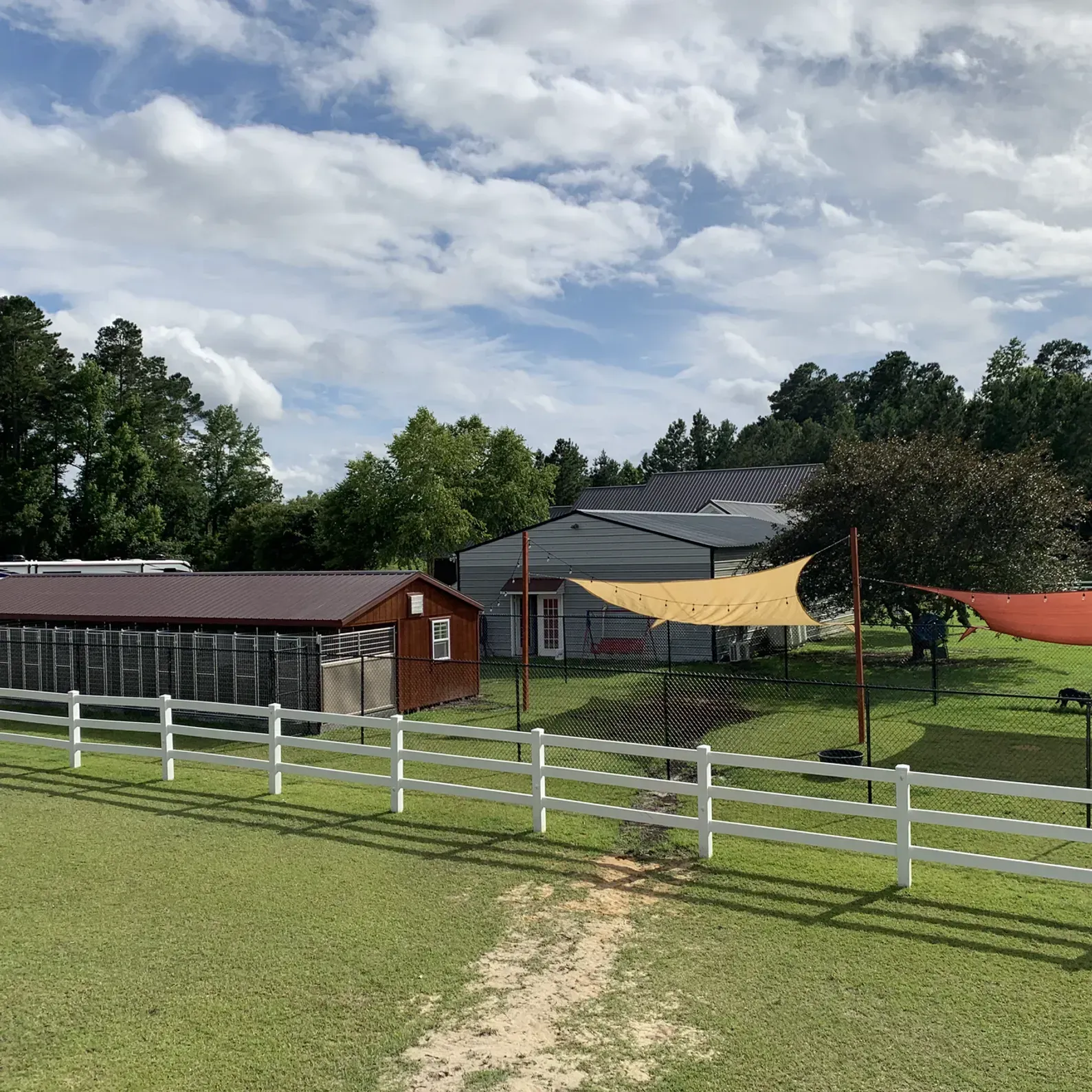 A rural scene: a white fence encloses a grassy area with a small house, shed, and shade sails under a cloudy sky.