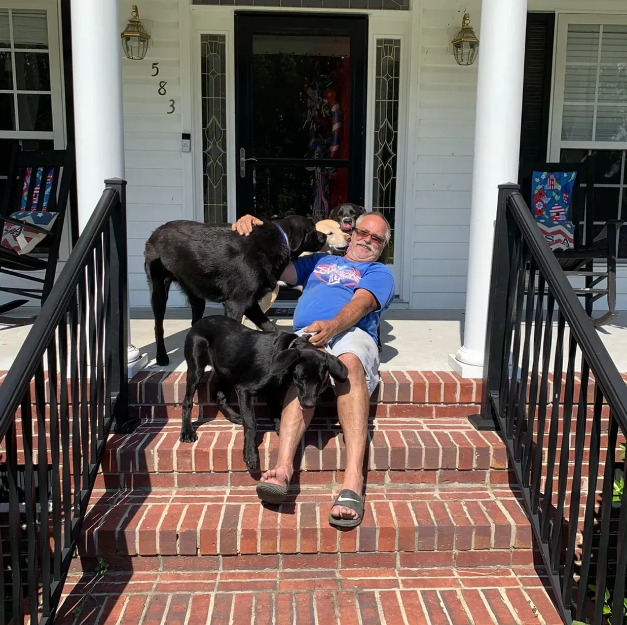 Man sitting on brick steps with three black dogs; house in background.