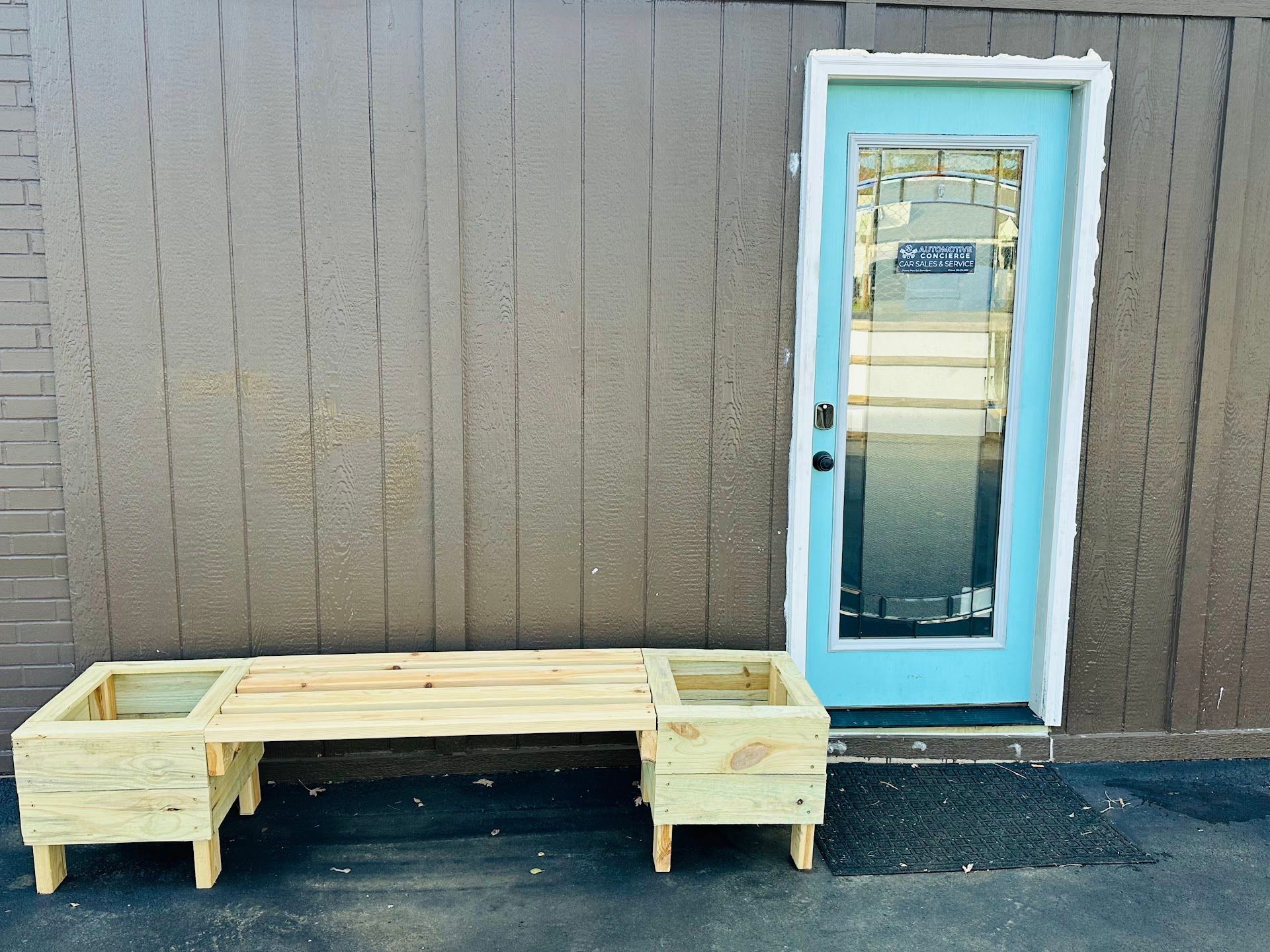 Wooden bench with planter boxes beside a blue-framed glass door on a brown building | Automotive Concierge KC
