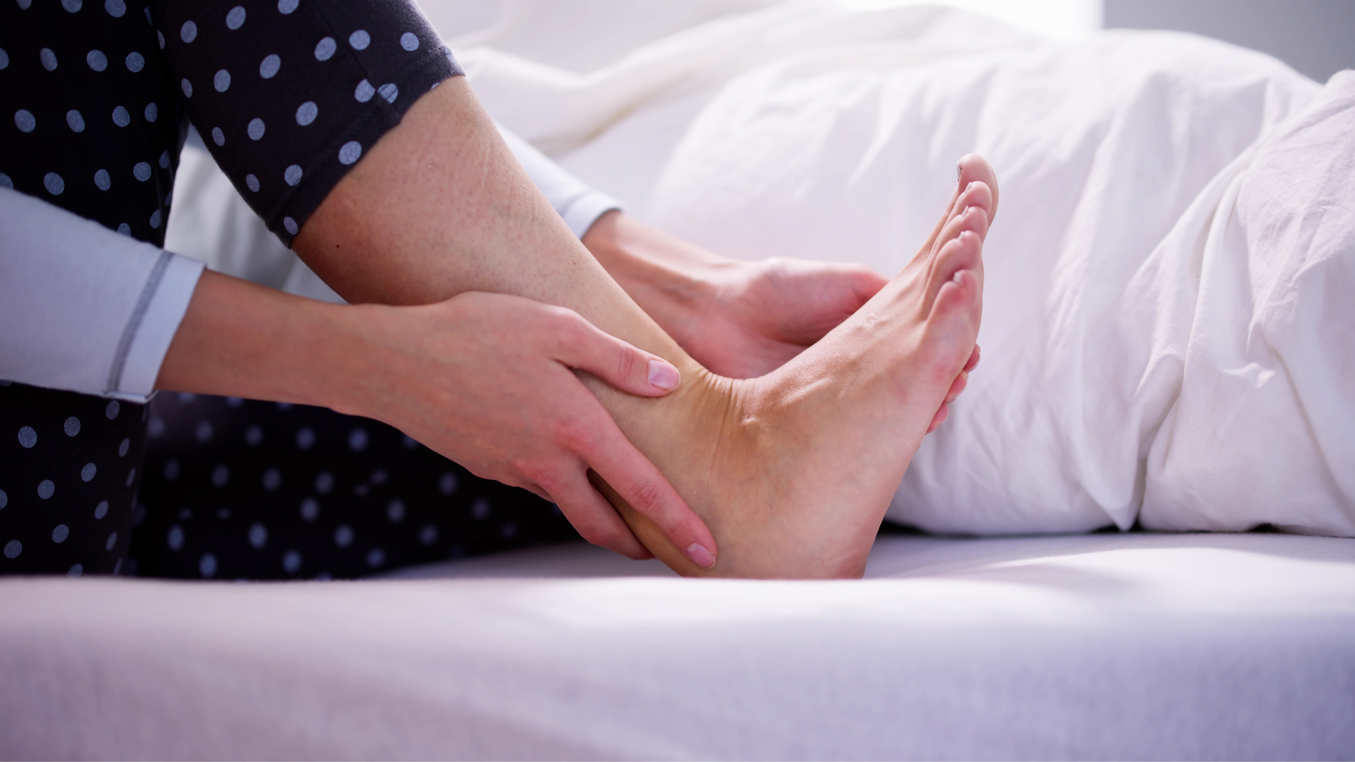 A woman is sitting on a bed holding her ankle in pain.