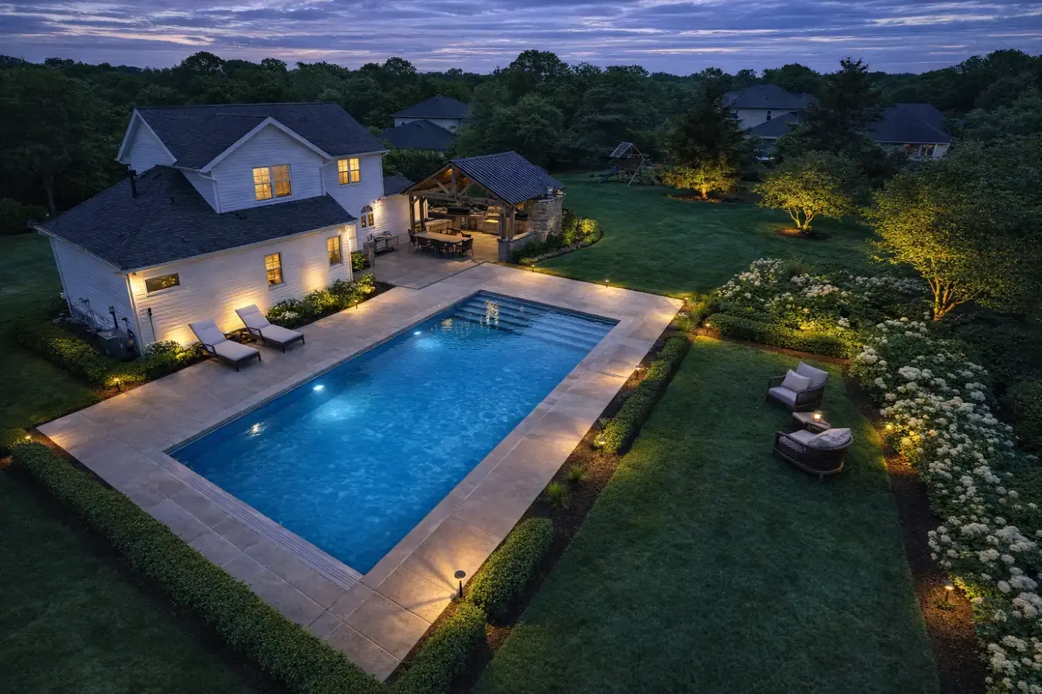 Backyard pool patio at dusk with lit umbrella, lounge chairs, and decorative lighting.