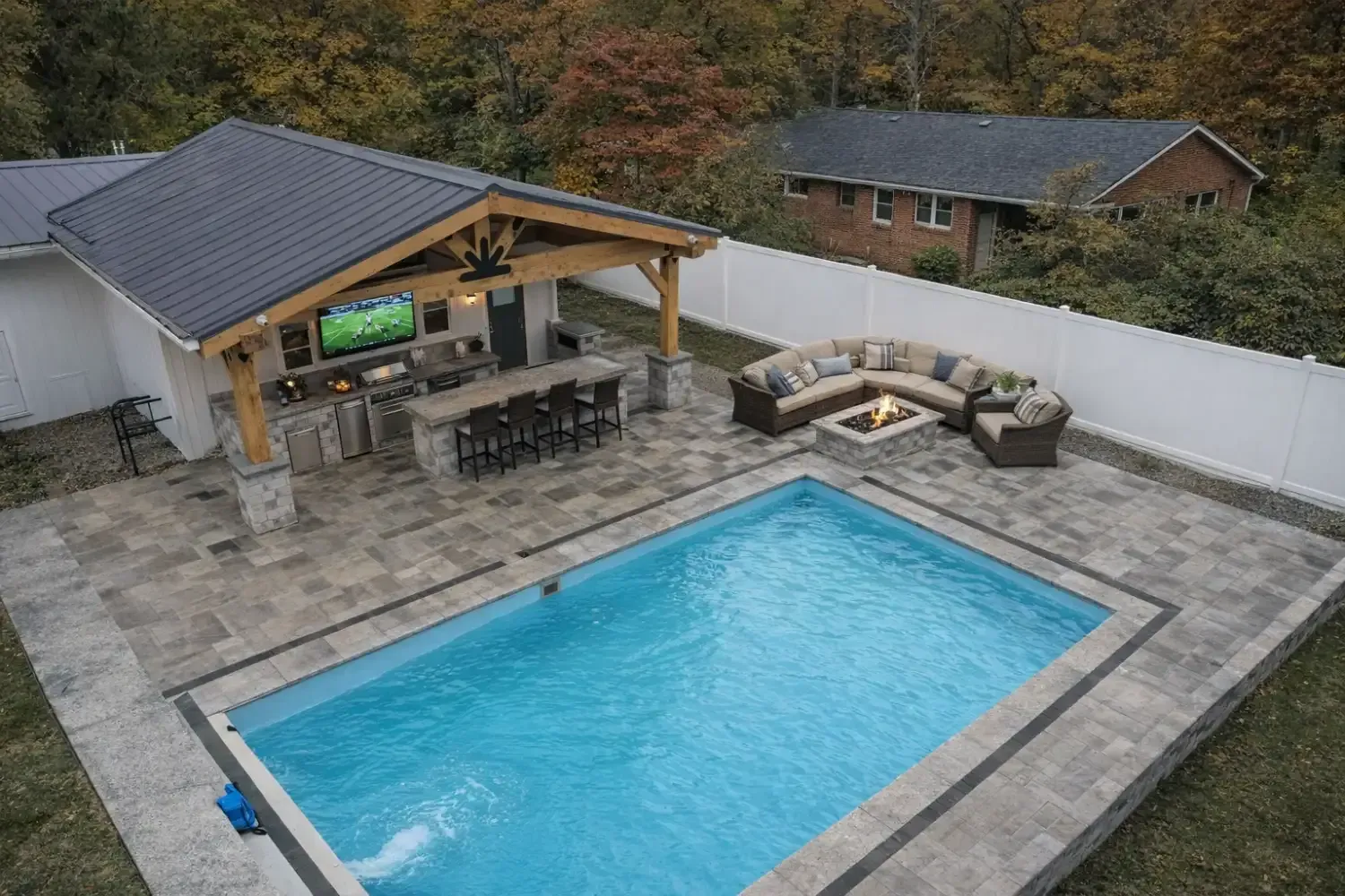Aerial view of a backyard with a rectangular swimming pool, stone patio, outdoor kitchen, and fire pit area.