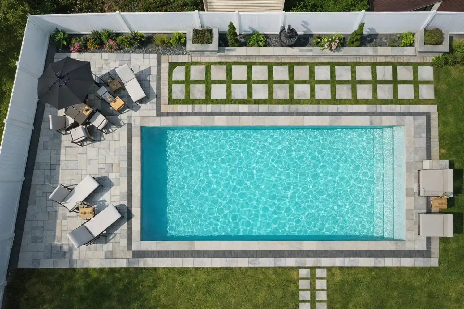 An aerial view of a rectangular backyard pool with light blue water, surrounding gray patio stones, and lounge seating.