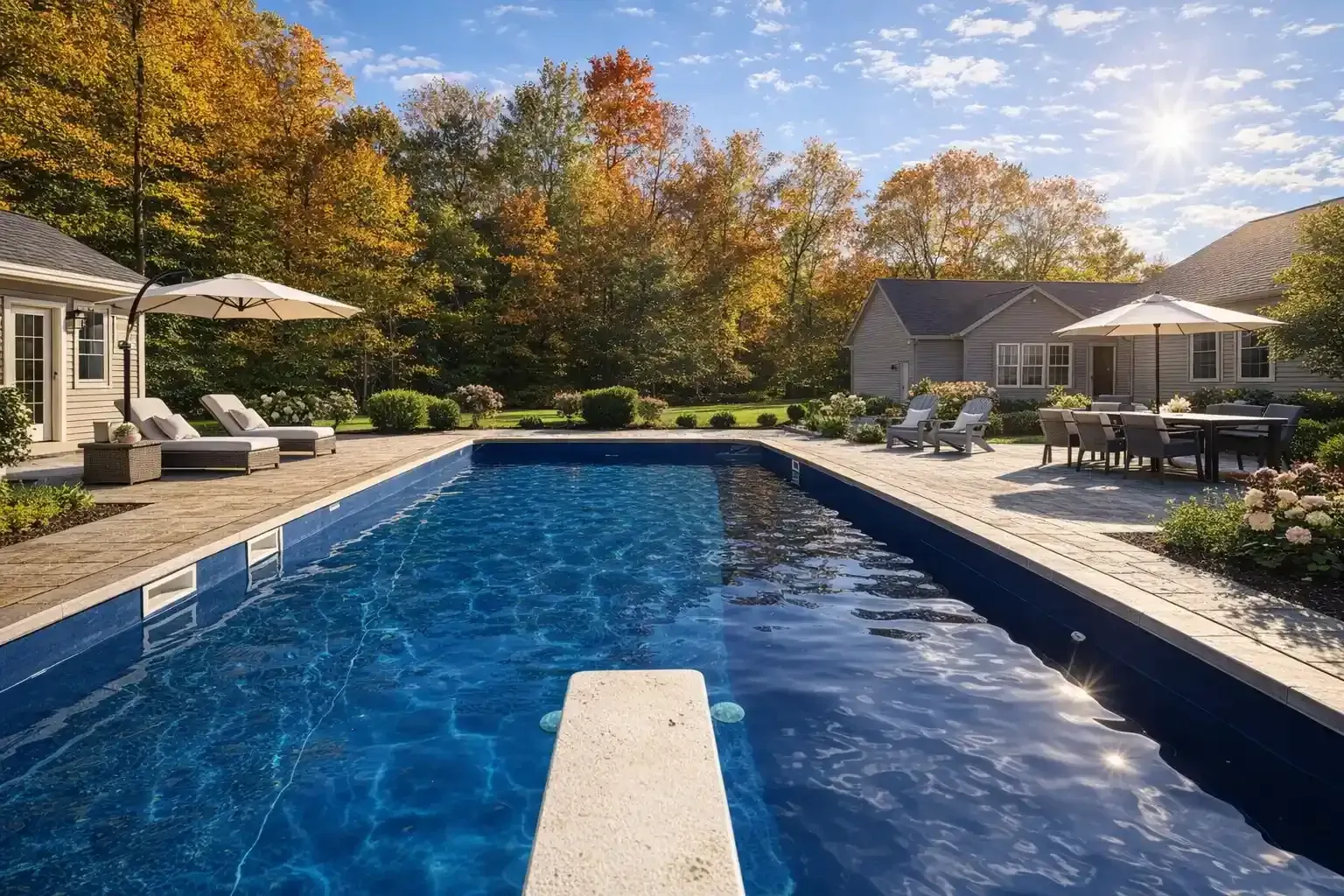 A rectangular swimming pool with a diving board, surrounded by patio furniture and autumn trees against a blue sky.