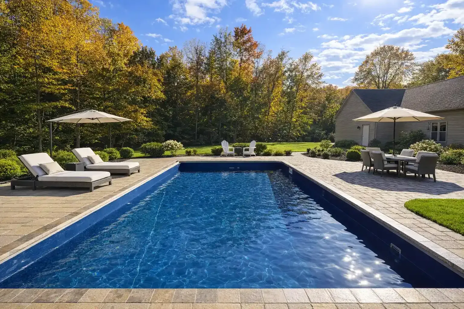 A rectangular swimming pool surrounded by a stone patio, lounge chairs, and an umbrella, with autumn trees in the back.