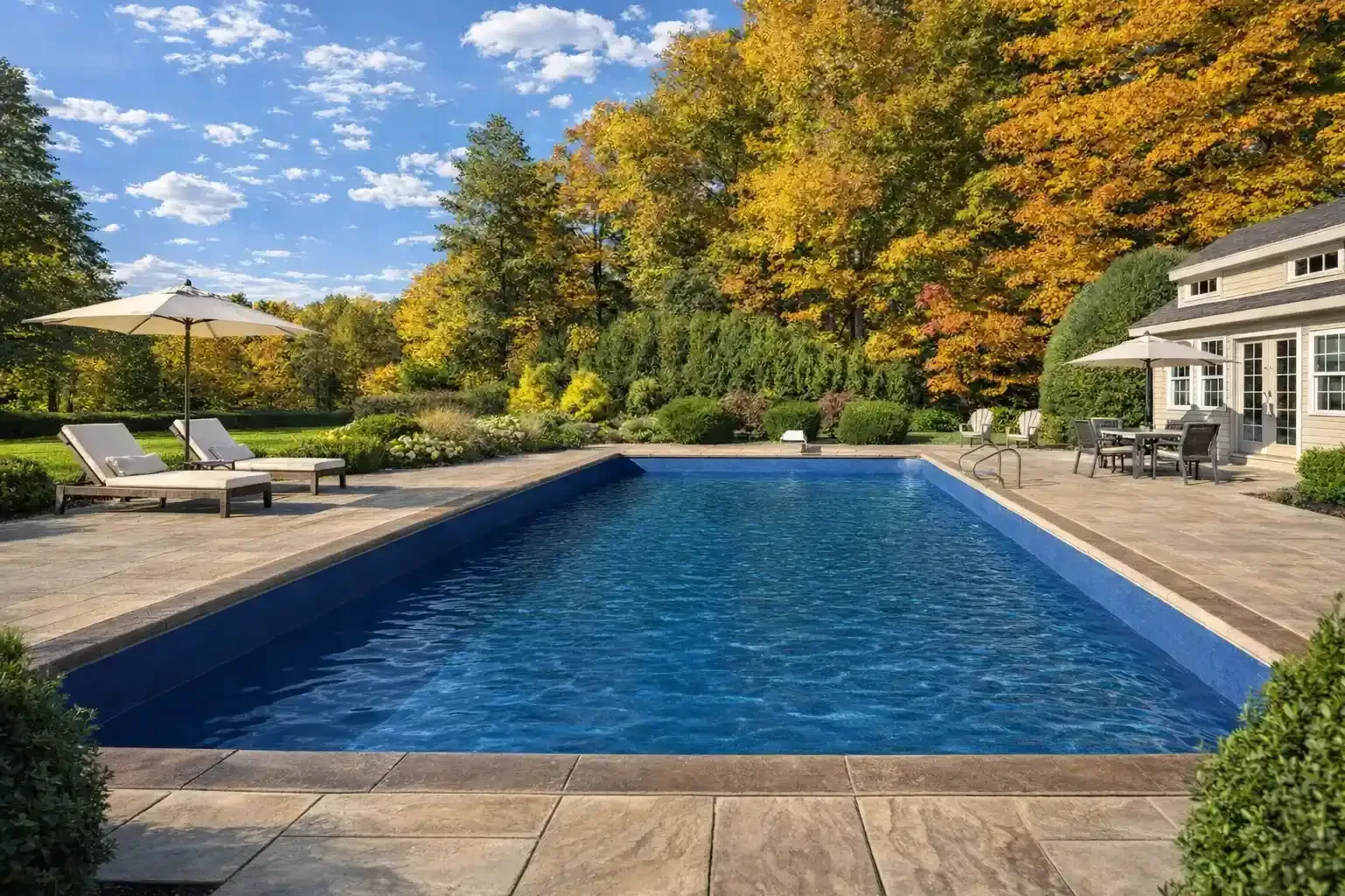 A rectangular swimming pool surrounded by a stone patio, loungers, and autumn trees under a sunny blue sky.