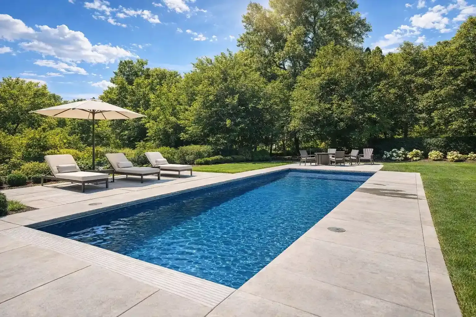 A rectangular blue swimming pool surrounded by a light-colored stone patio, lounge chairs, and a lush green treeline.