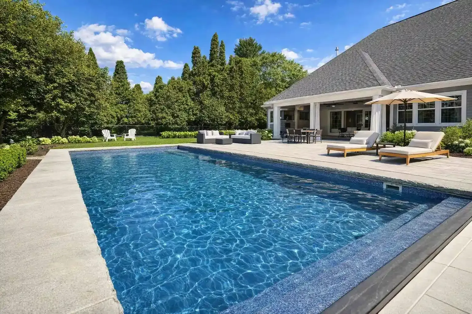 An outdoor swimming pool with bright blue water, surrounded by a stone patio, lounge chairs, and lush green trees.