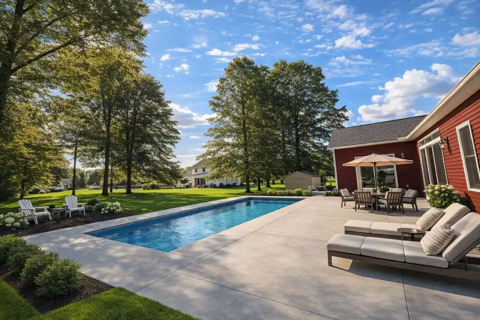 A rectangular swimming pool set in a backyard with a red house, patio furniture, lounge chairs, and large green trees.