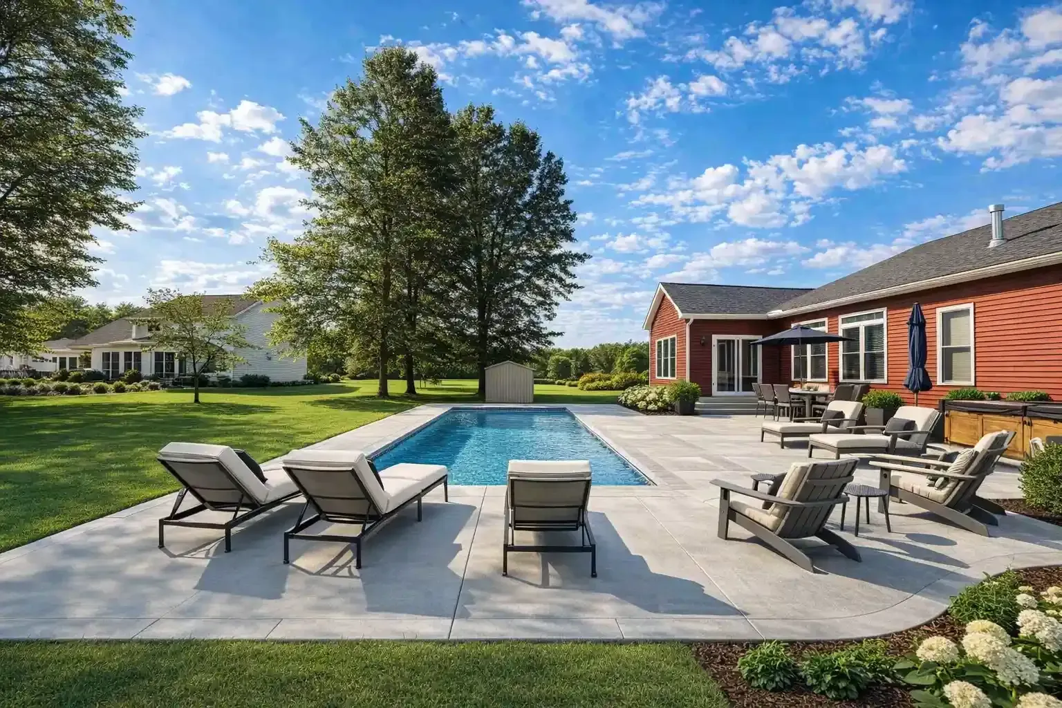 A swimming pool surrounded by a patio and lounge chairs in a sunny, grassy backyard beside a large red house.