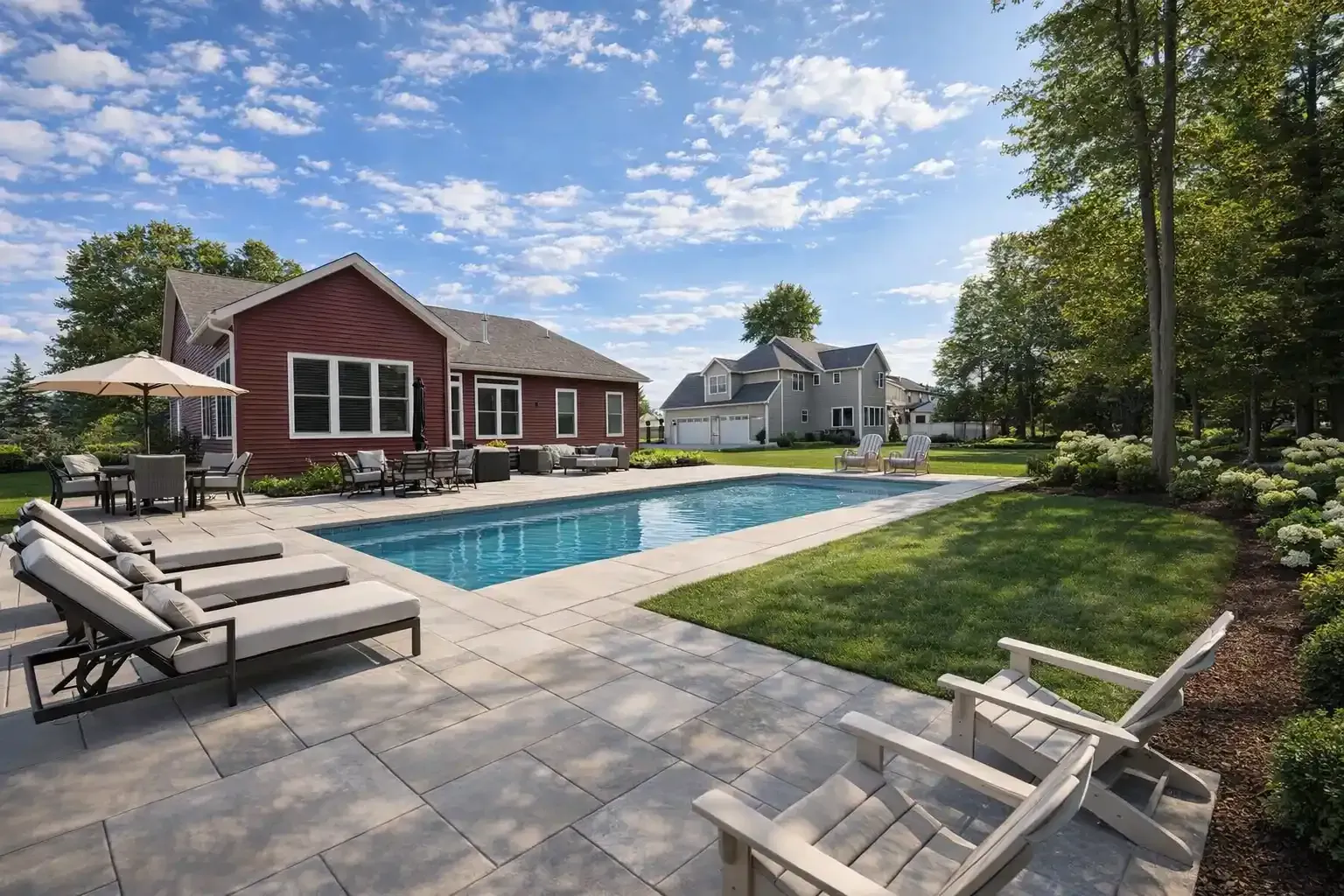 A rectangular swimming pool in a stone patio backyard with lounge chairs, a dining set, and a red house under blue sky.