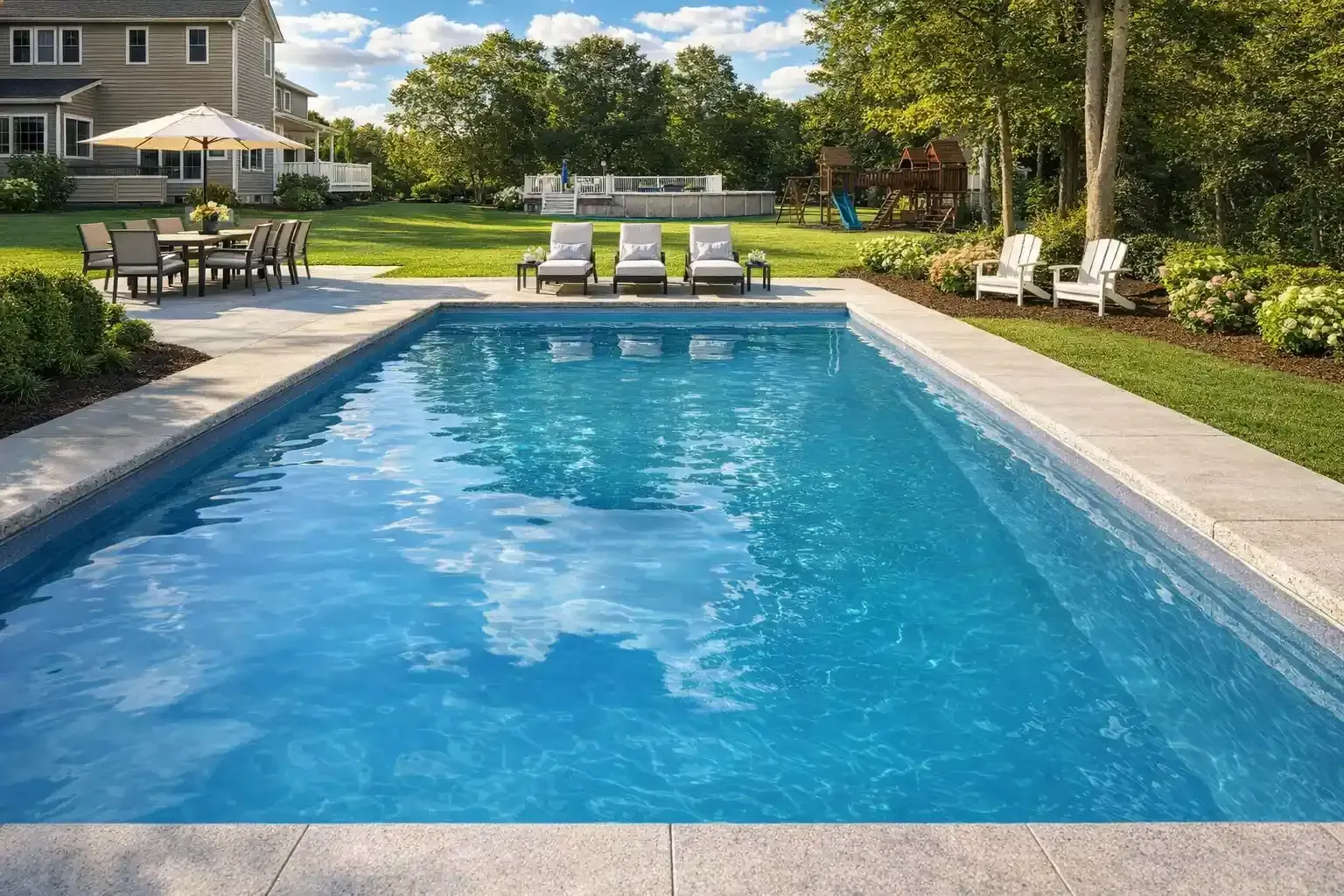 A rectangular backyard swimming pool with blue water, surrounded by patio furniture, a lawn, and trees on a sunny day.