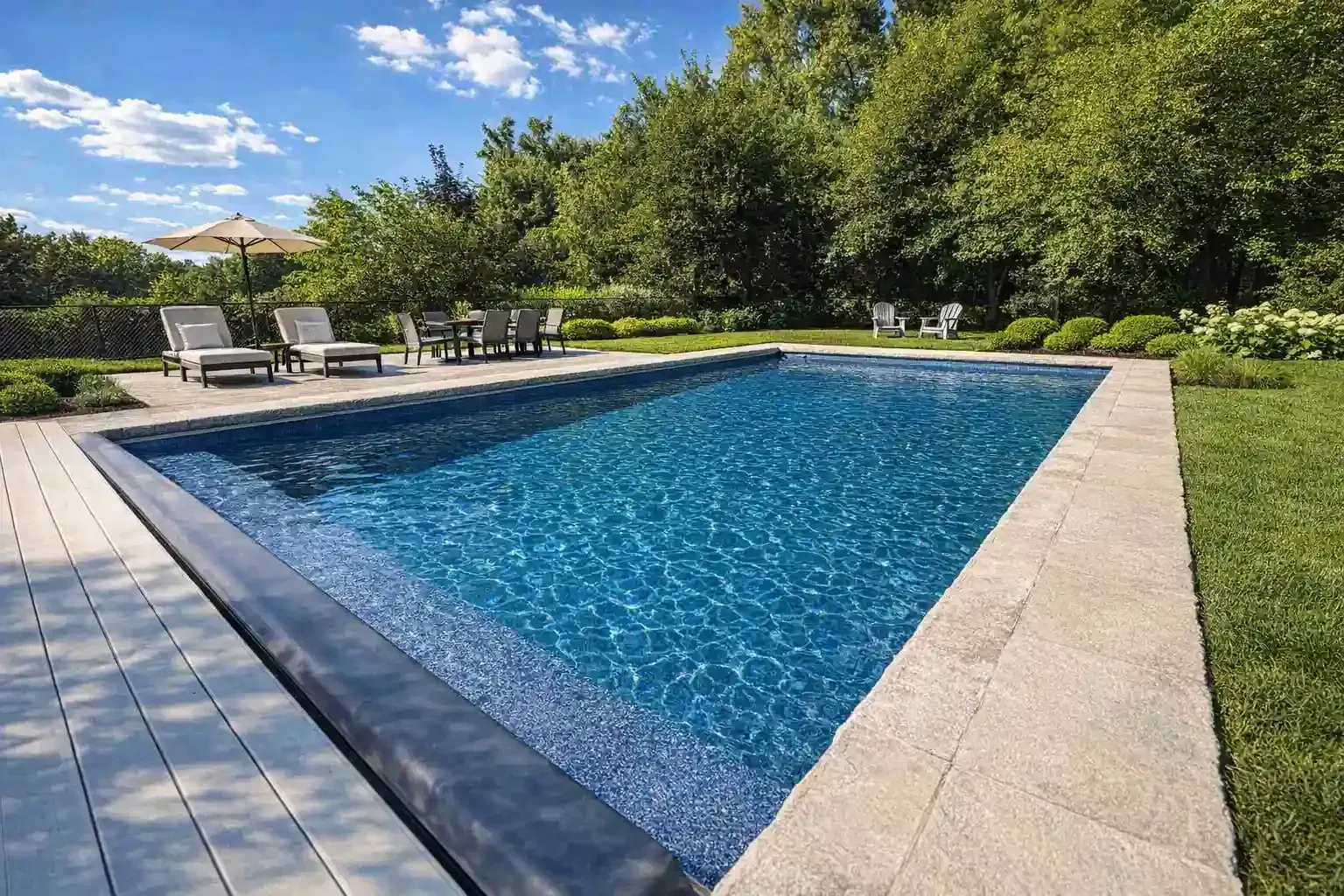 A rectangular swimming pool with deep blue water, surrounded by stone decking, lounge chairs, and lush green trees.