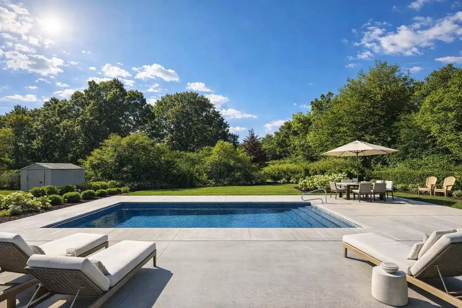 A rectangular swimming pool surrounded by a concrete patio, lounge chairs, and an umbrella, set against lush green trees.