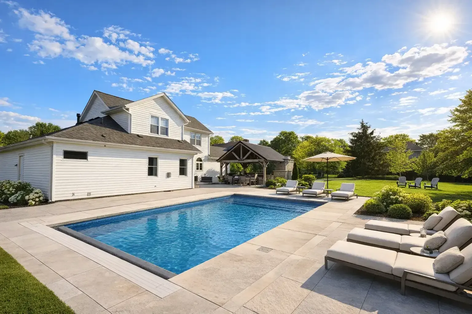 A white two-story house with a rectangular pool, stone patio, lounge chairs, and a gazebo on a sunny, grassy lawn.