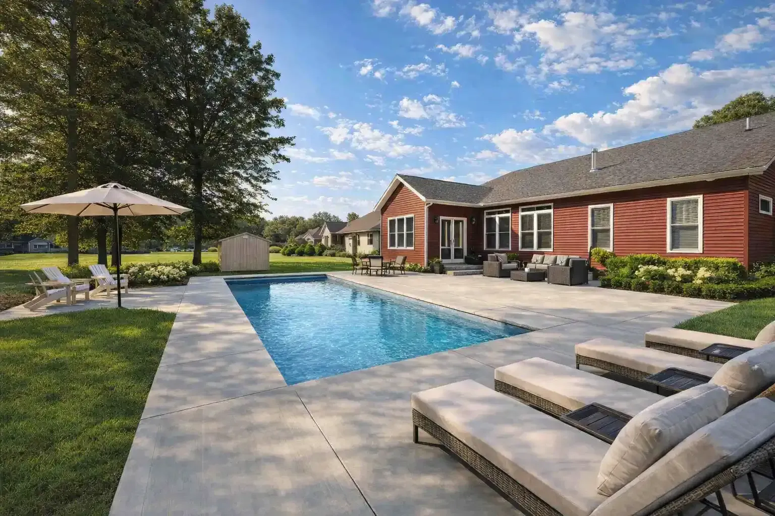 A rectangular swimming pool on a concrete patio beside a red house with lounge chairs and a patio umbrella on a sunny day.