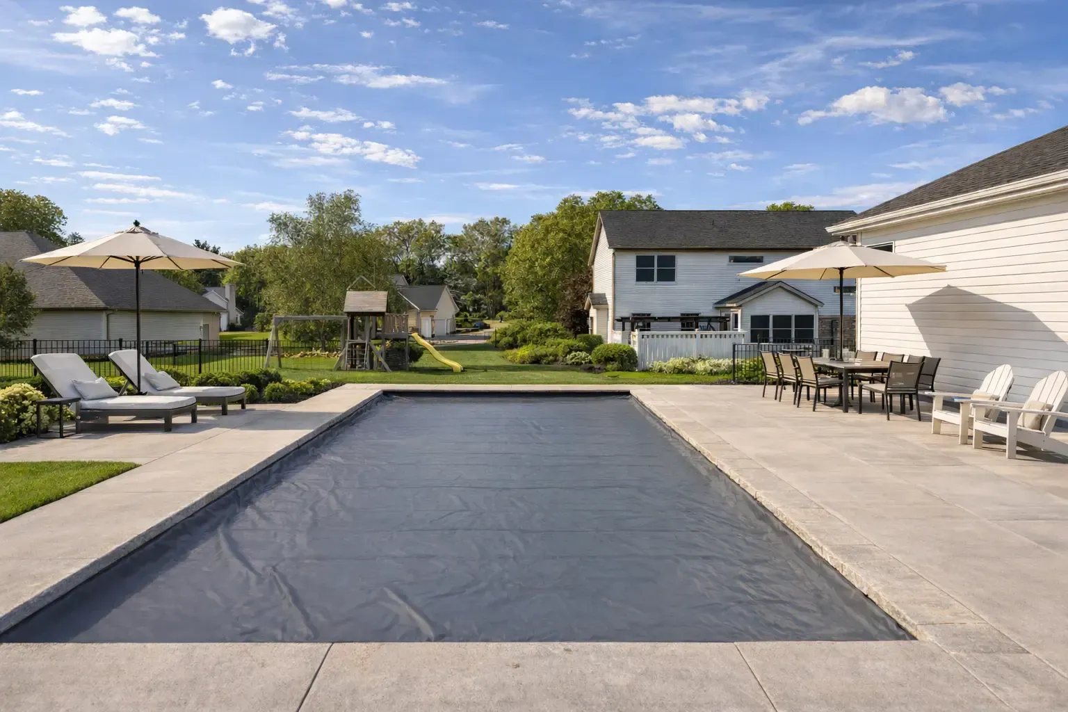A rectangular swimming pool covered with a dark tarp on a sunny patio, surrounded by lounge chairs and umbrellas.