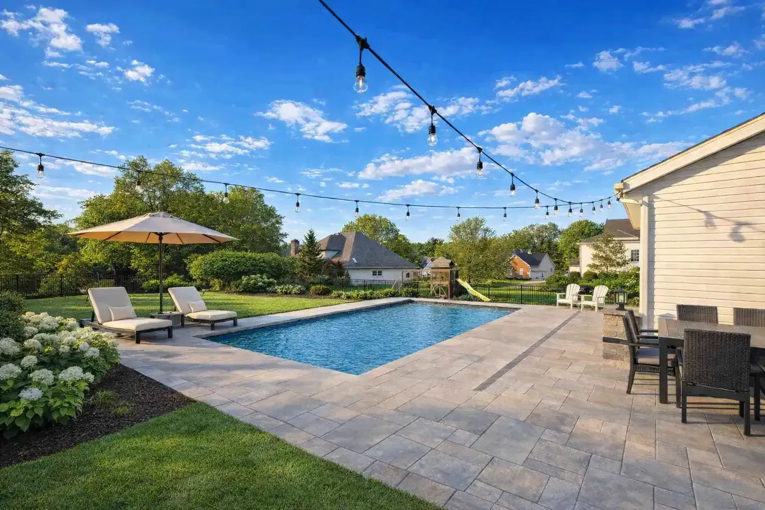 A backyard swimming pool with lounge chairs, an umbrella, and string lights on a sunny day next to a house patio.