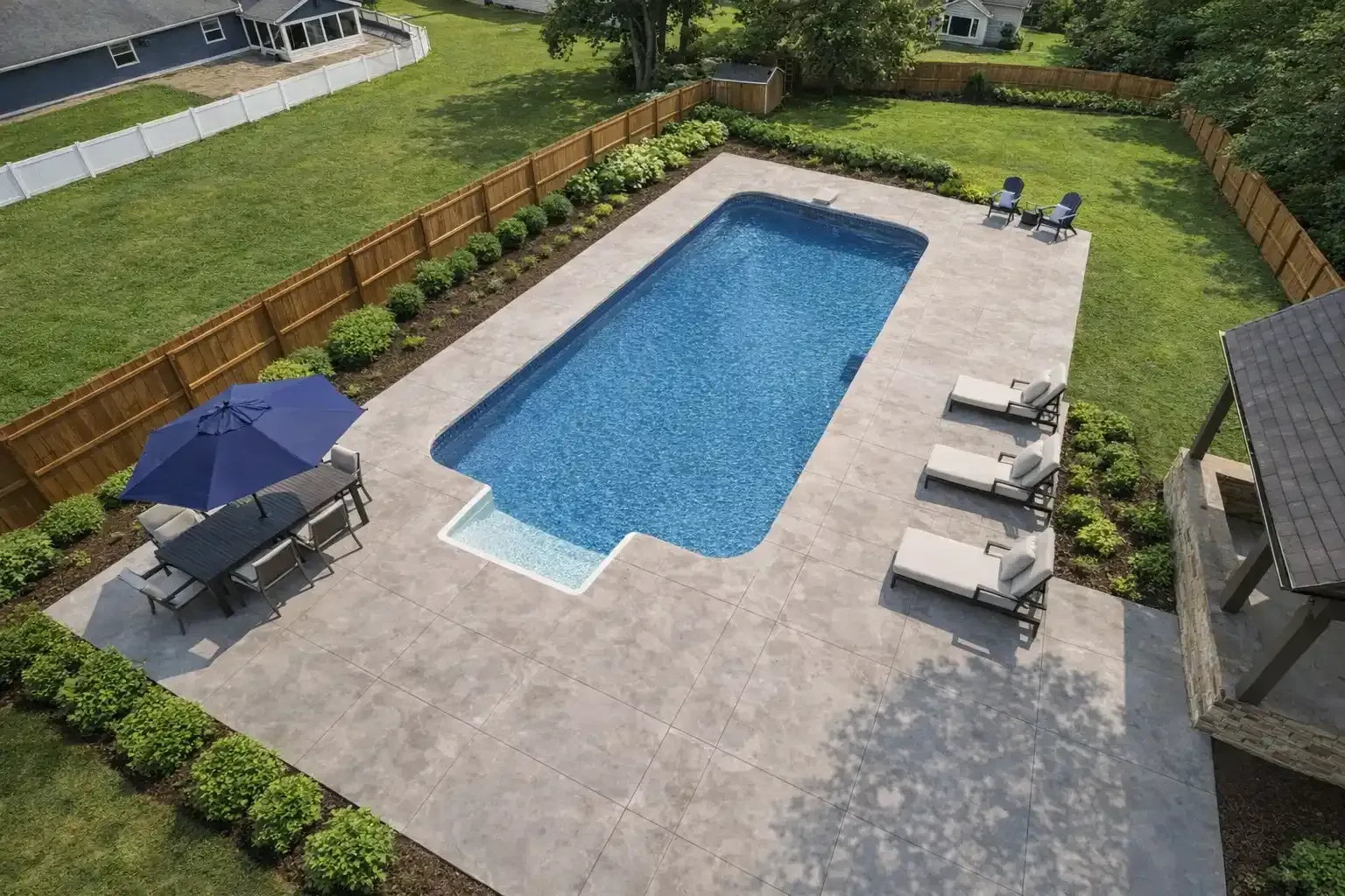 An aerial view of a backyard swimming pool with a blue umbrella, dining set, and lounge chairs on a stone patio.