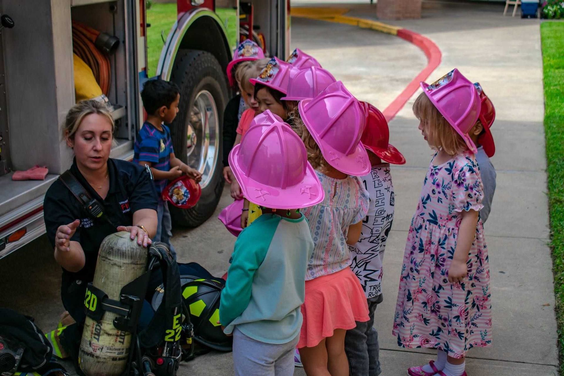 A group of children wearing pink firefighter hats are standing in front of a fire truck.