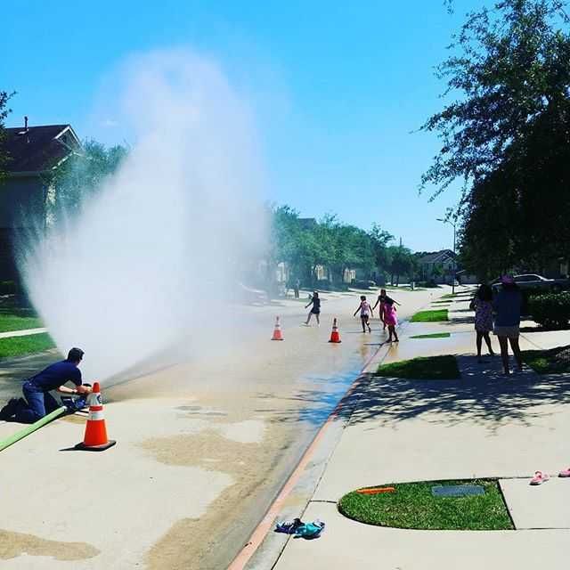 A firefighter is spraying water from a hose on a sidewalk and kids are playing in the water.