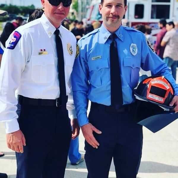 Two men in uniform pose for a picture with one holding a helmet that says firefighter