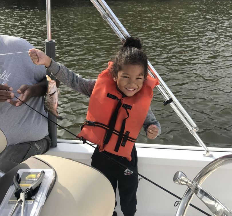 A little girl wearing an orange life vest is fishing on a boat