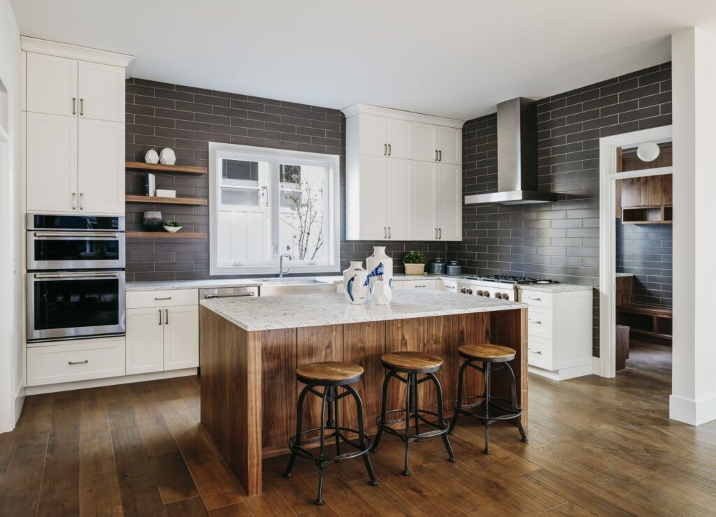 A modern kitchen with white upper cabinets, a wooden island with three stools, and a dark tiled backsplash wall.