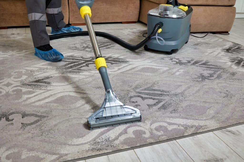 A worker using an extraction machine to deep clean a patterned carpet in a room with a sofa.