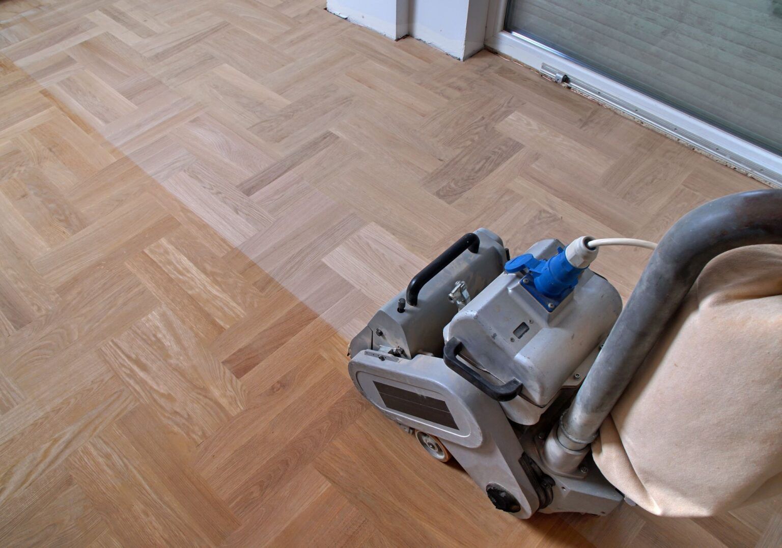 A drum sander refinishing a light-colored herringbone hardwood floor in a room.