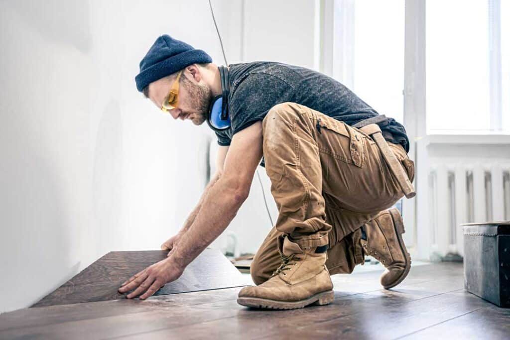 A construction worker wearing a beanie and safety glasses kneeling to install brown laminate flooring in a bright room.