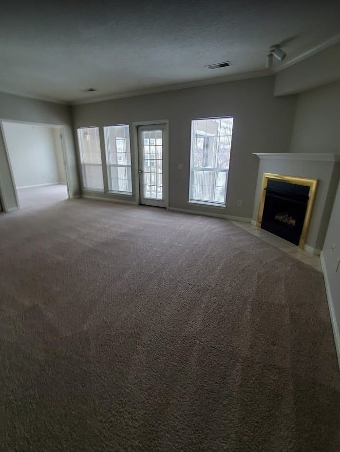 An empty living room with tan carpet, neutral walls, large windows, a door, and a fireplace with a gold frame.