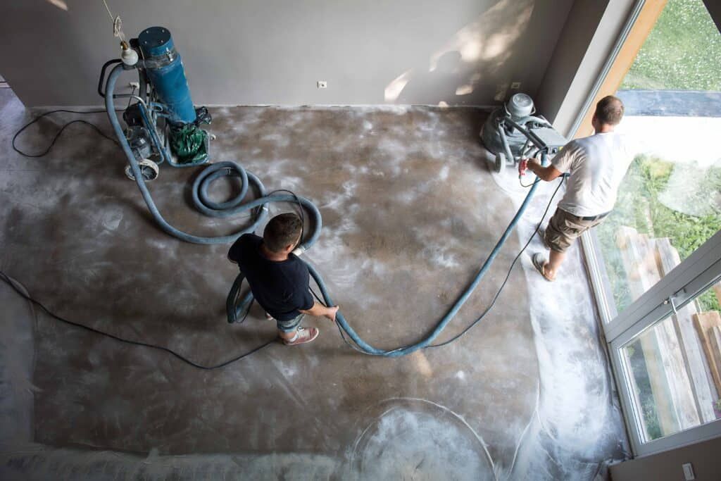 Two people in a room use industrial grinding equipment and hoses to polish a concrete floor.