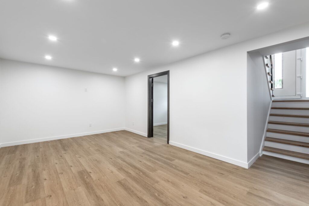 An empty, minimalist basement room with white walls, recessed ceiling lights, light wood flooring, and wooden stairs.