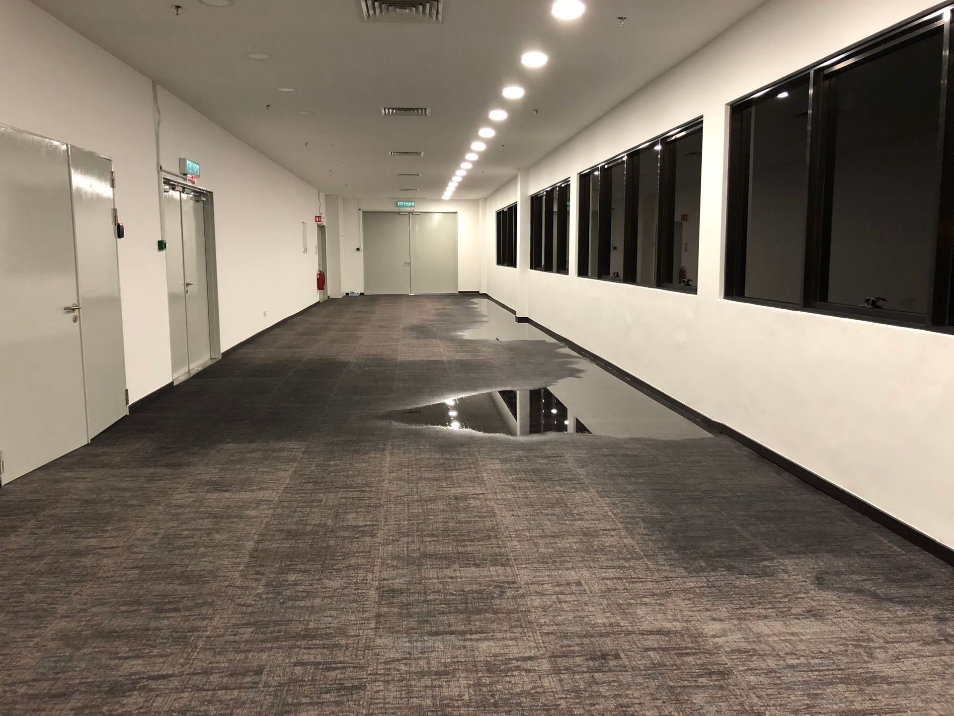A long, narrow office hallway with white walls and dark carpet featuring a large, reflective water spill on the floor.