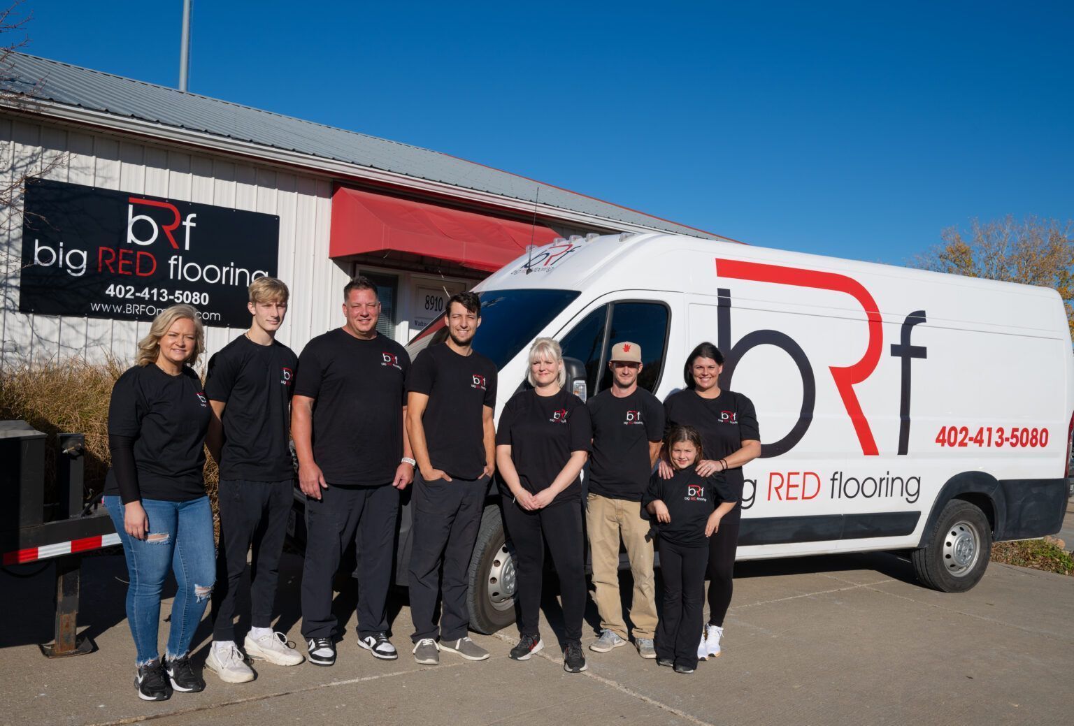 A group of people in black shirts stands in front of a Big Red Flooring work van parked outside their office.
