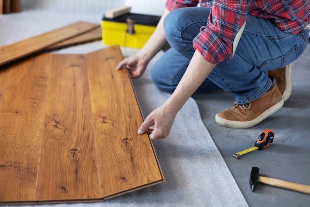 A person in jeans and a red plaid shirt installs wood-look laminate flooring panels on a gray underlayment.