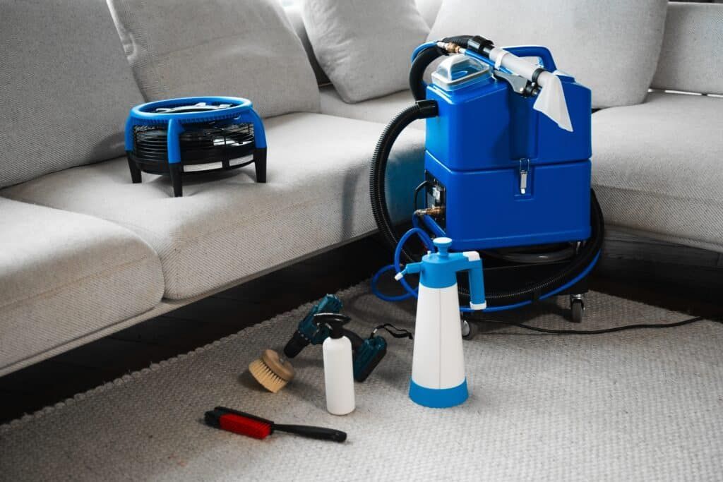 Professional upholstery cleaning equipment and various tools arranged on a carpet in front of a white sofa.