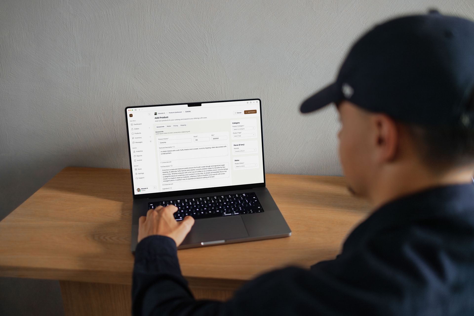 Person wearing a blue cap typing on a laptop at a wooden desk, indoors. The laptop screen displays data.