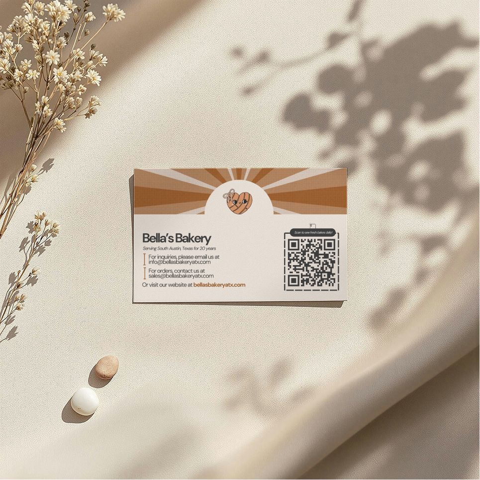 Business card: Bella's Bakery, on a cream satin cloth with shadows, dried flowers, and two small candies.