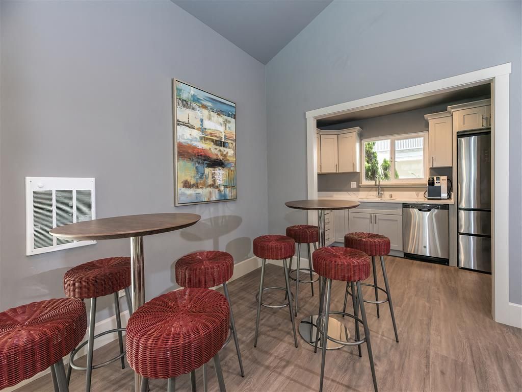 Common lounge with round wooden tables and red woven stools; kitchenette visible in background.
