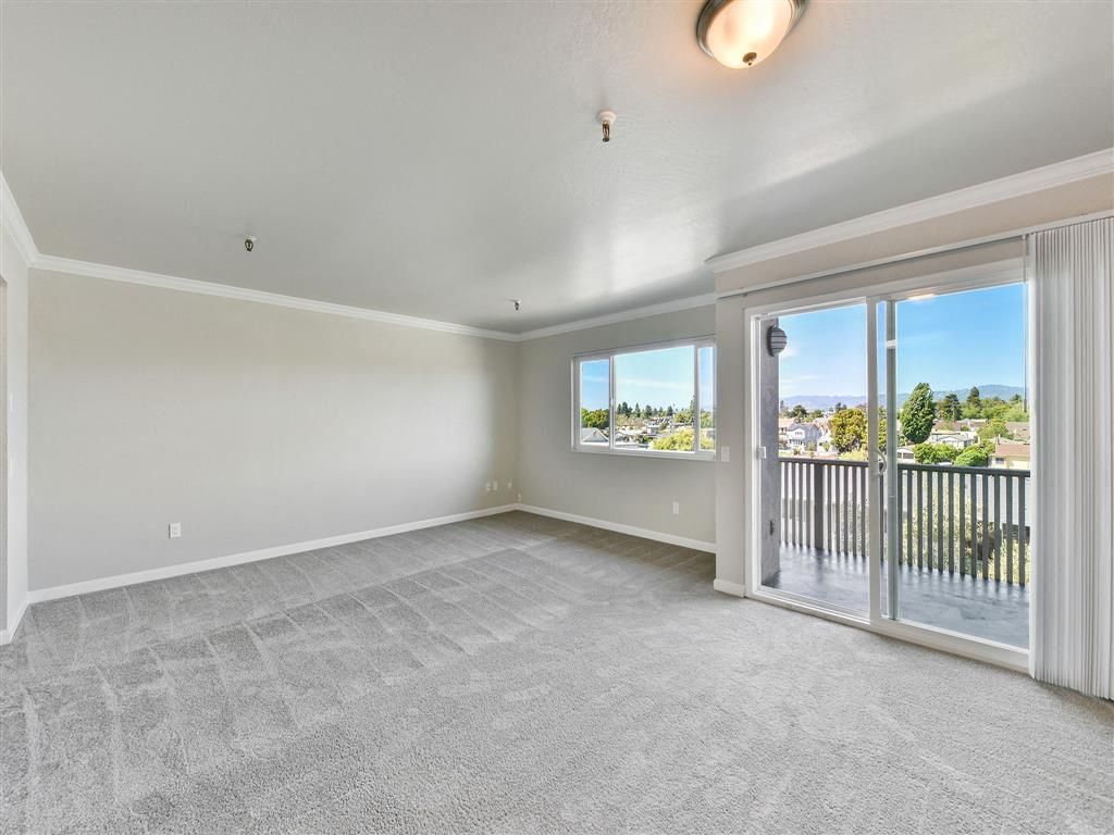 Bright empty living room with beige walls, gray carpet, and a sliding glass door to a balcony.