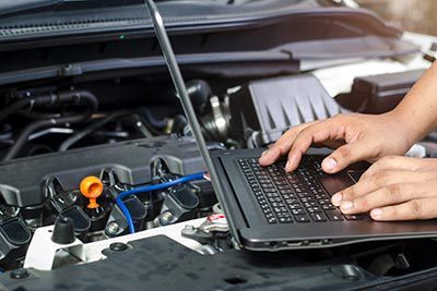 A person is working on a laptop computer under the hood of a car.