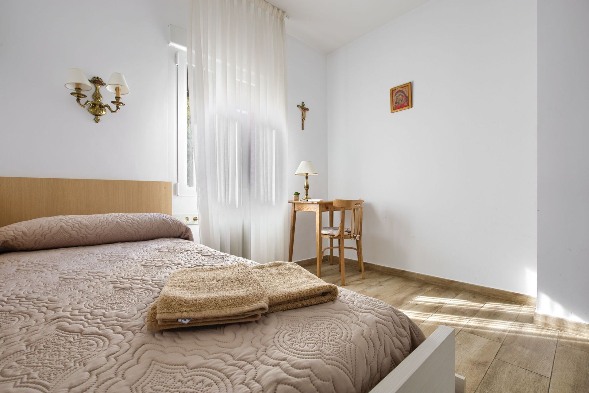Bedroom with a bed, small table, and sheer curtains. Light brown and white color scheme.