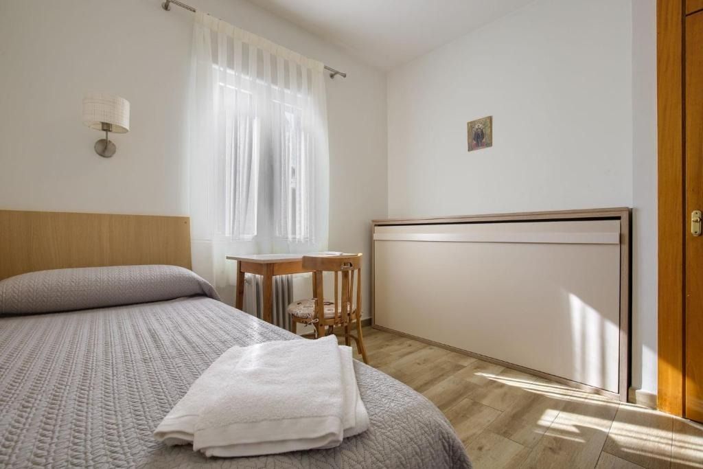 Small, simply furnished bedroom with a bed, small table, and a window with sheer curtains.