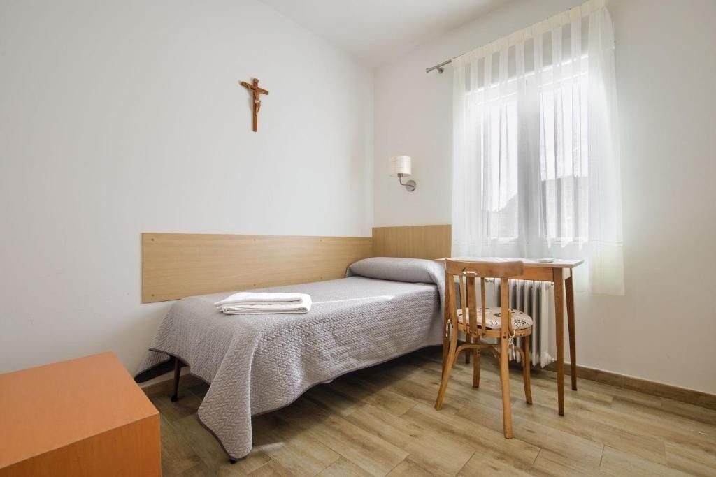 Bedroom with single bed, small table and chair, crucifix, and window with curtains.