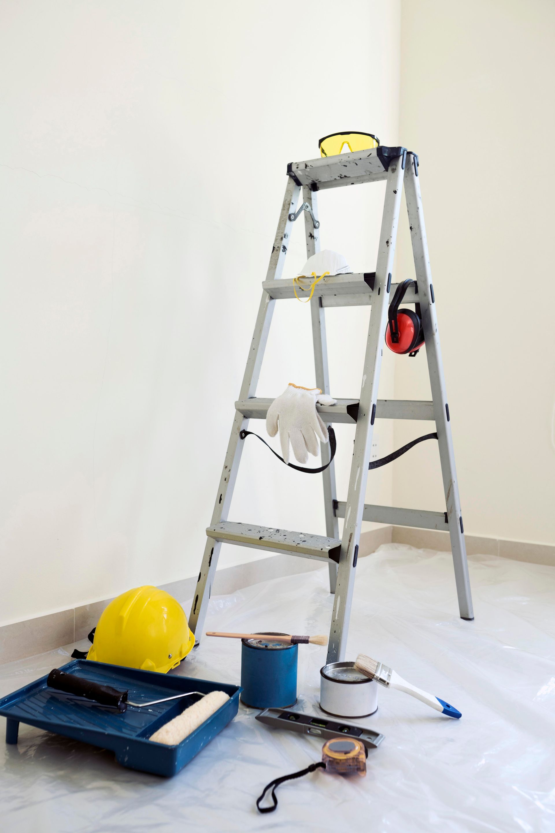 A ladder is sitting on the floor next to a hard hat and paint supplies.