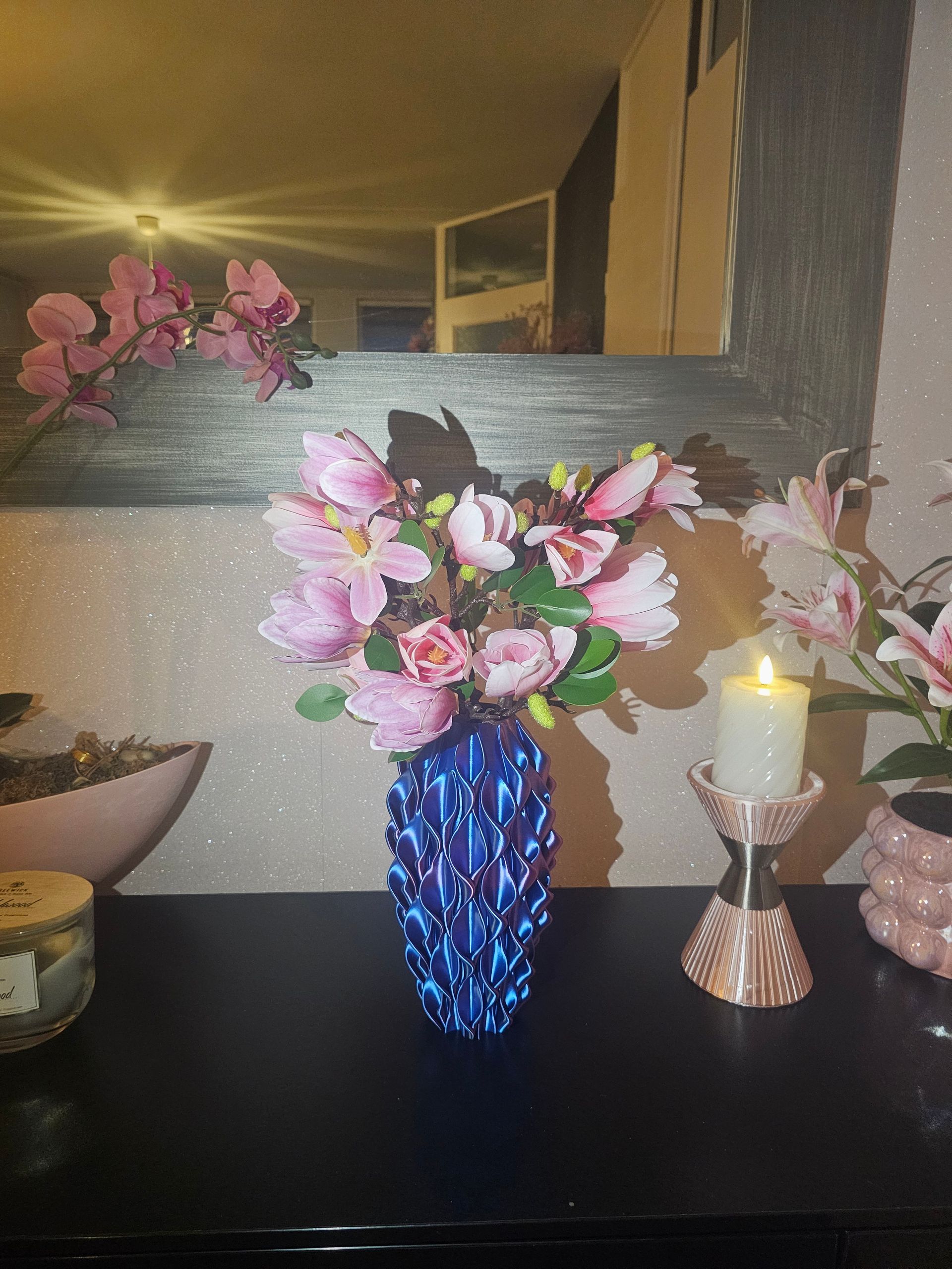 Blue beaded vase holds pink and purple flowers on a black surface; a lit candle and mirror in the background.