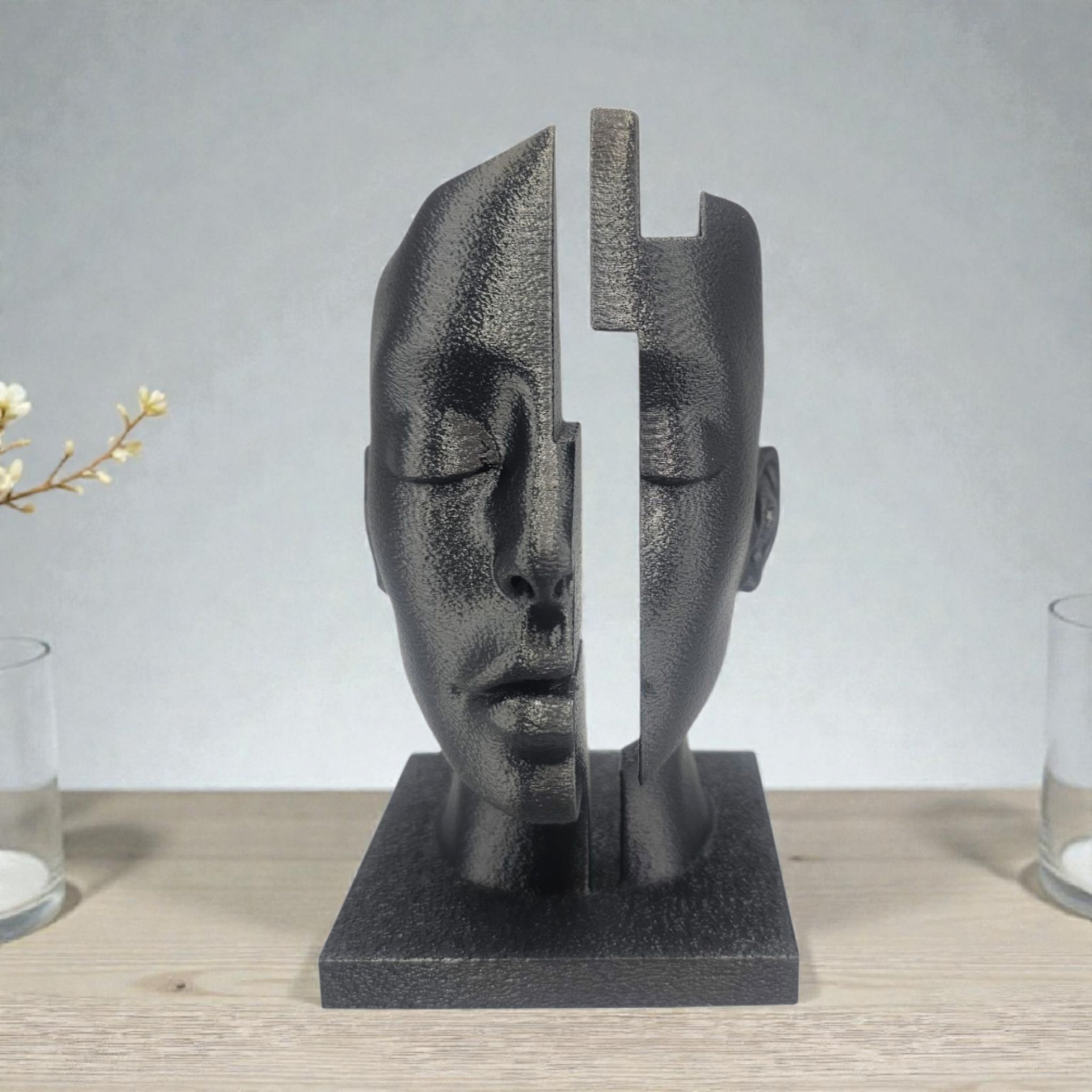 Zwarte sculptuur van een in tweeën gesplitst gezicht, met een kruisvormige holte in het midden; op een houten ondergrond.